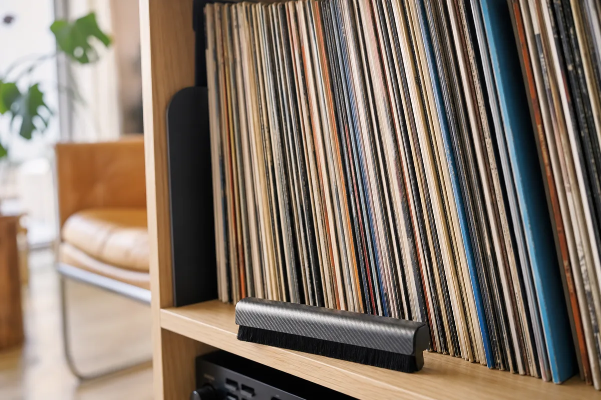 Disques vinyles rangés verticalement sur une étagère en bois clair, une brosse antistatique posée devant