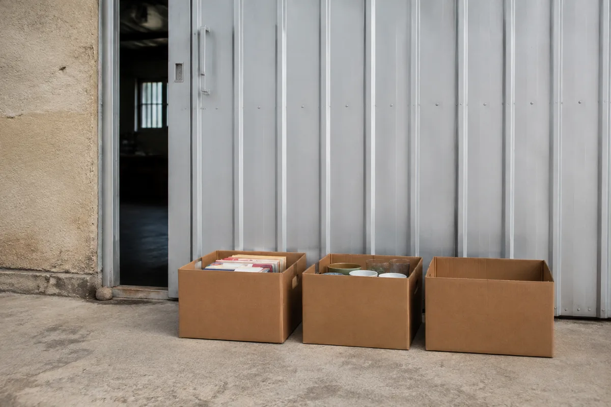 Cartons bruns remplis de livres et de vaisselle posés devant une porte de hangar en tôle claire